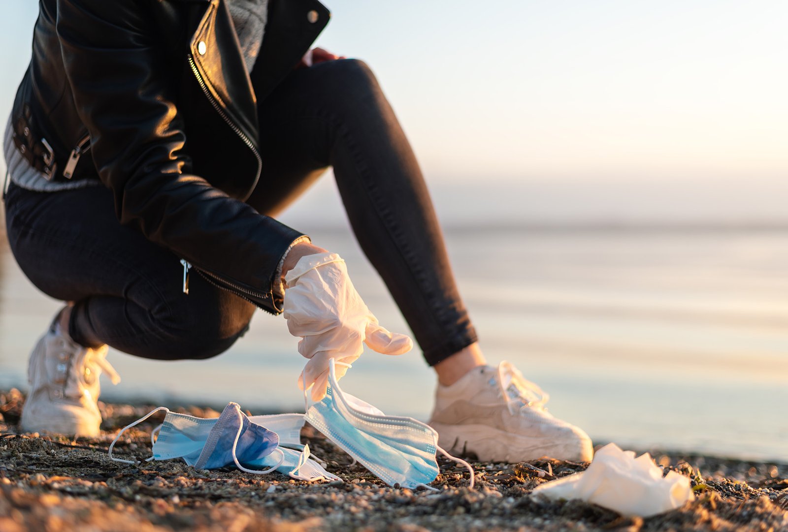Young woman picking up disposable medical mask on beach. Post co